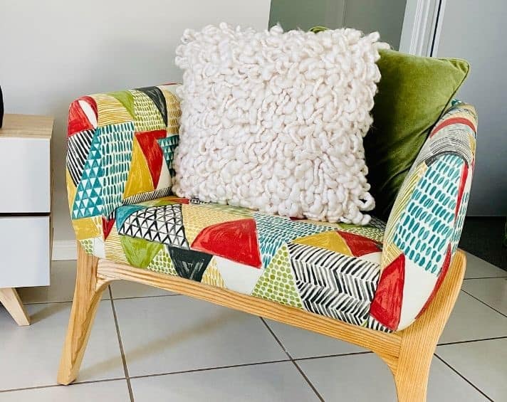 Colourful patterned armchair with wooden legs, featuring a white textured cushion and a green cushion, placed on a tiled floor next to a piece of light wood furniture.