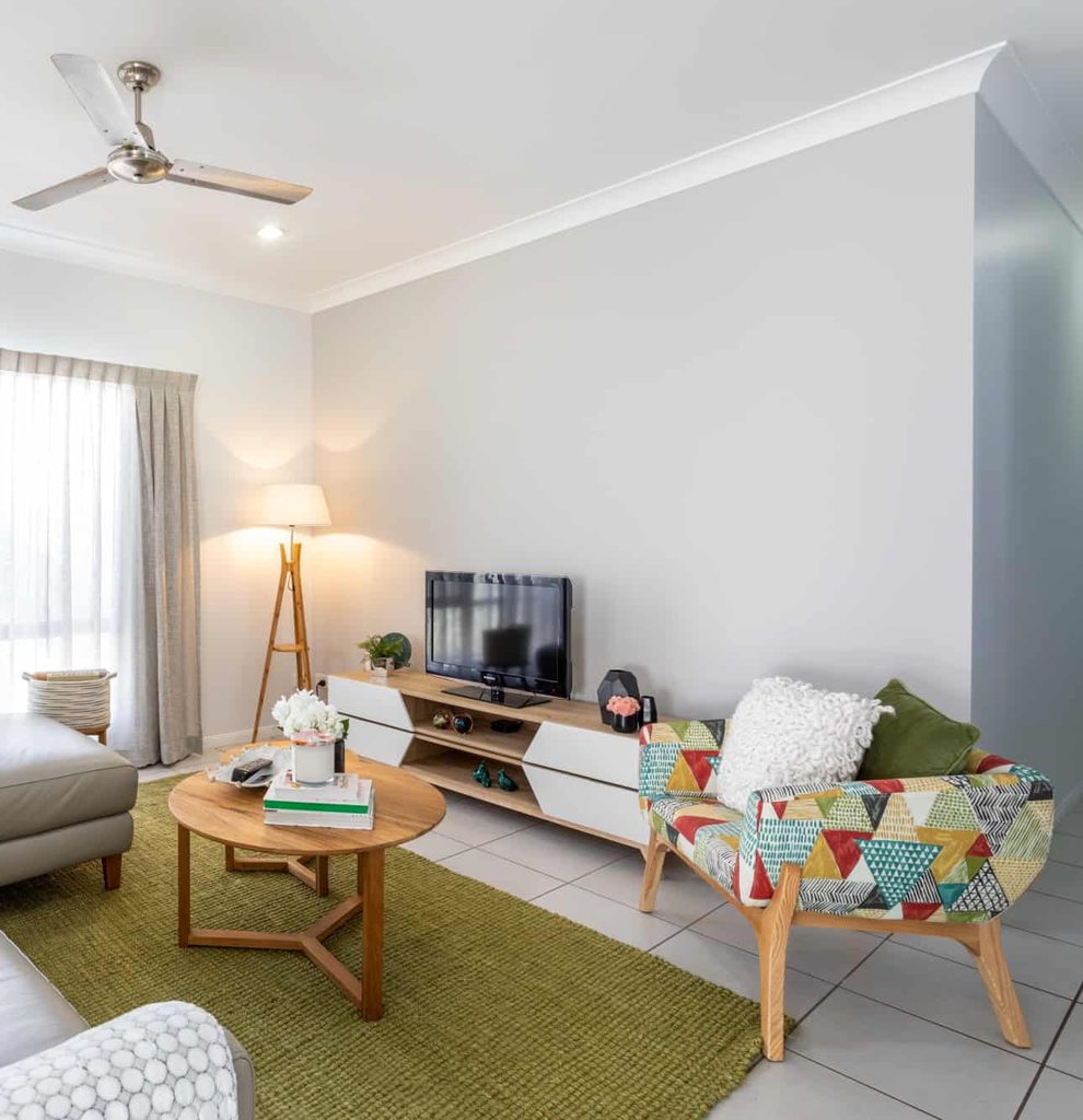 Modern living room with a TV on a low cabinet, colourful armchair, round wooden coffee table, green rug, floor lamp, and light grey walls.
