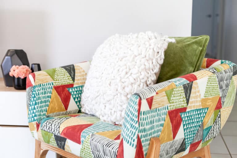 Colourful patterned armchair with a green and a chunky white cushion, next to a small table holding a black vase and pink flowers.