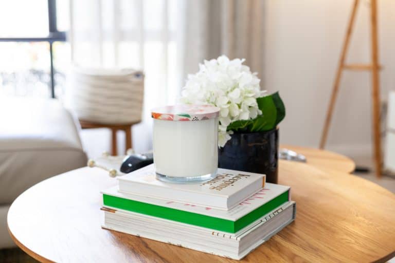 A glass candle sits on a stack of books next to a black vase with white flowers on a wooden table in a bright living room.