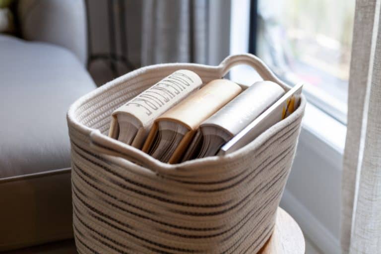 A woven basket with stripes holds several hardback books, placed on a small wooden table near a window with net curtains.