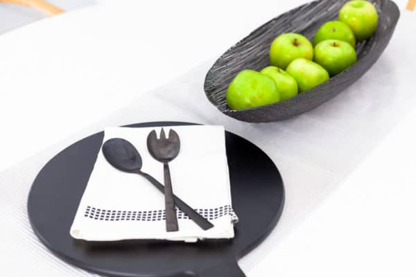 Black spoon and spork on a folded white serviette next to a black bowl filled with green apples, all placed on a white table.
