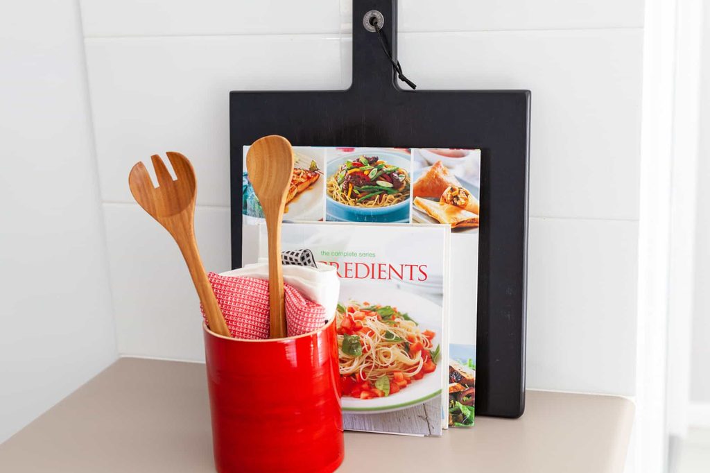 A red ceramic container holds wooden utensils and a cloth in front of a cookery book and a black chopping board on a kitchen worktop.