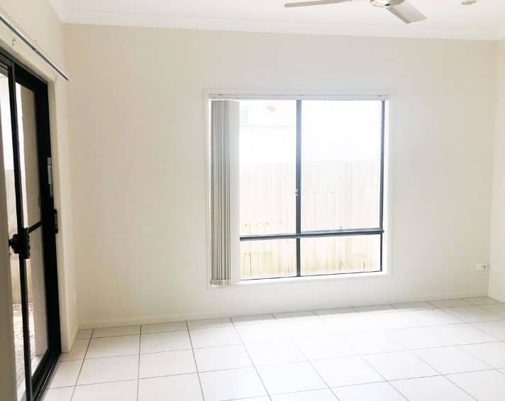Bright empty room with white tiled floor, large window with vertical blinds, sliding glass door on the left, and a ceiling fan.