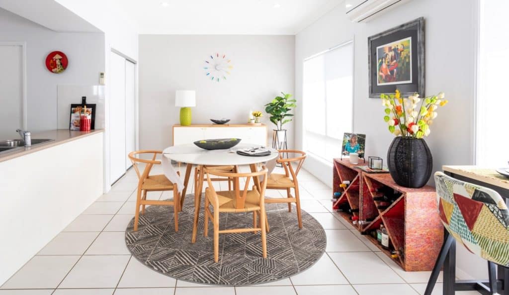 Modern dining room with a round table, four wooden chairs, decorative shelves, wall art, and bright natural light.