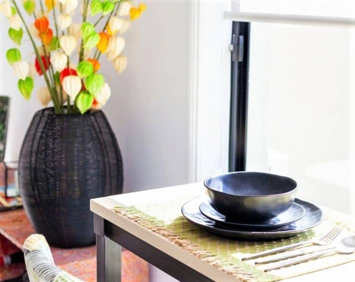 Black ceramic bowl and plate with cutlery set on a table near a window, with a black vase holding colourful artificial leaves in the background.