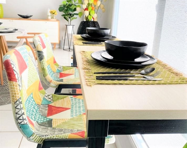 Two colourful chairs at a dining table set with black crockery, placemats, and cutlery. Bright, modern interior with plants and natural light.