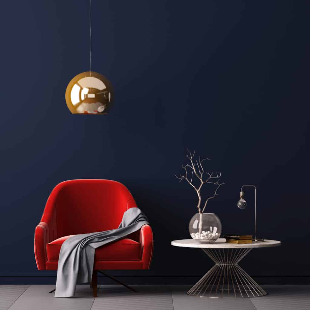 A red armchair with a grey blanket sits beside a round white table with a decorative branch, fishbowl, books, and lamp, against a dark blue wall and gold hanging light fitting.