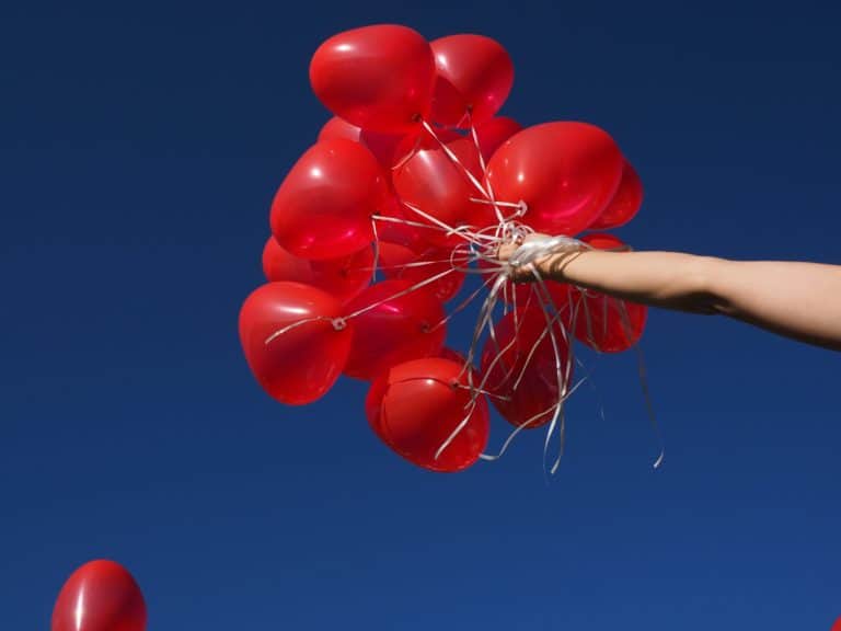 A person’s outstretched arm holds a bunch of red balloons against a clear blue sky.