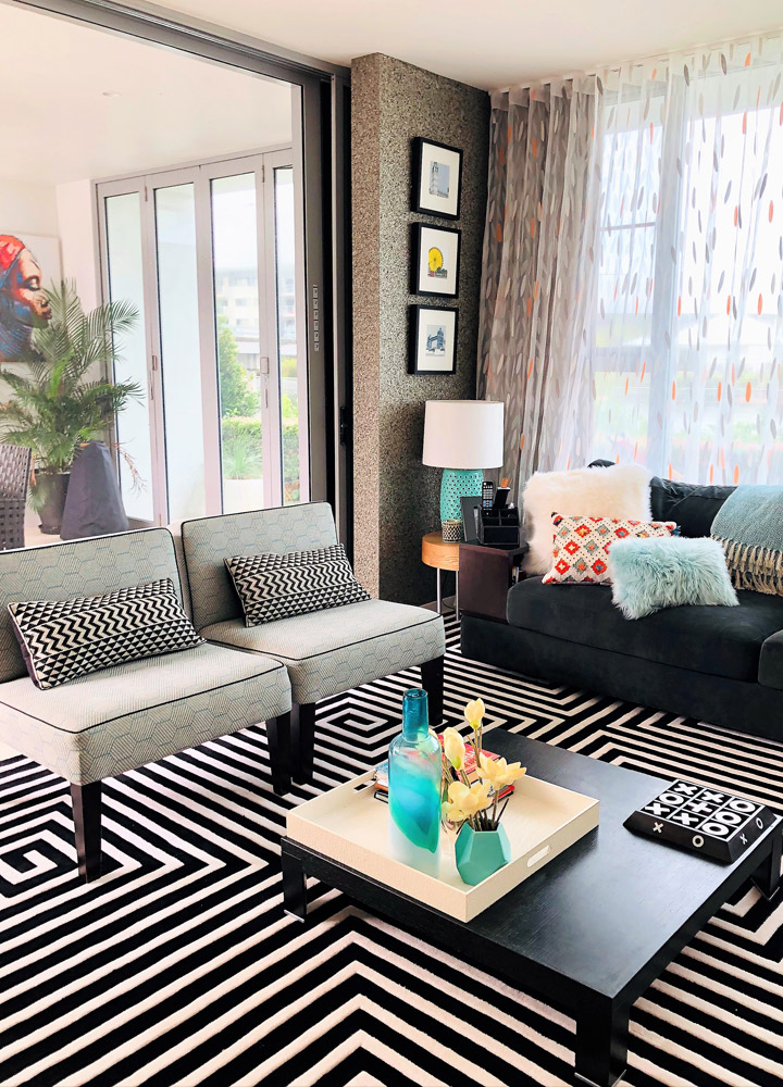 Modern living room with patterned rug, grey chairs, black sofa with colourful cushions, and a coffee table holding vases and books; large windows with sheer curtains and framed art on the wall.