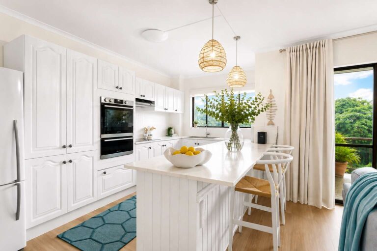 Bright kitchen with white cupboards, hanging lights, a bowl of lemons on the island, bar stools, a blue rug, and a large window with beige curtains overlooking greenery.