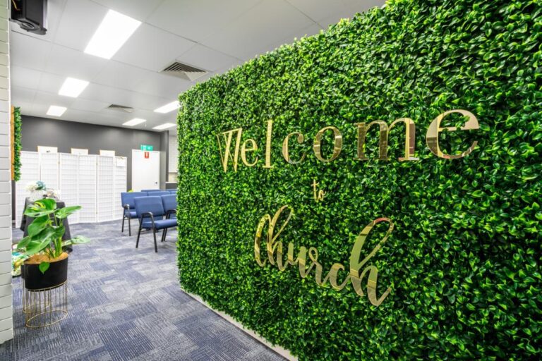 Indoor space with a green artificial hedge wall displaying the words “Welcome to Church” in gold lettering; blue chairs and white walls are visible in the background.