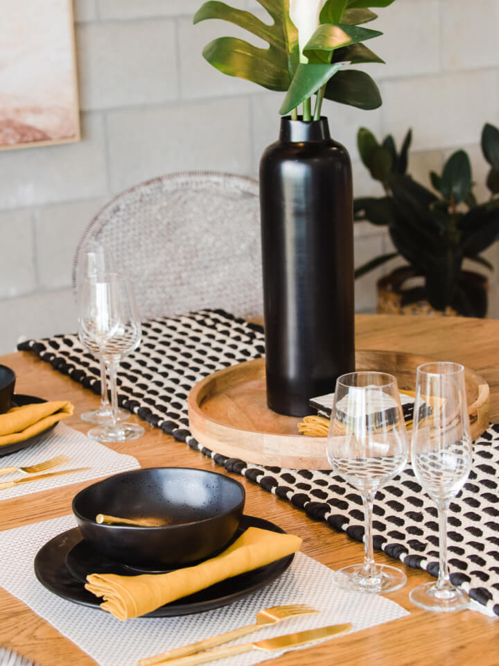 A wooden dining table is set with black crockery, yellow serviettes, wine glasses, and a tall black vase with flowers on a tray.
