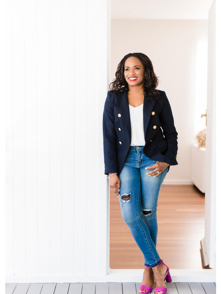 Woman standing in a doorway, wearing a navy blazer, white top, ripped blue jeans, and pink heels, smiling and looking to the side.