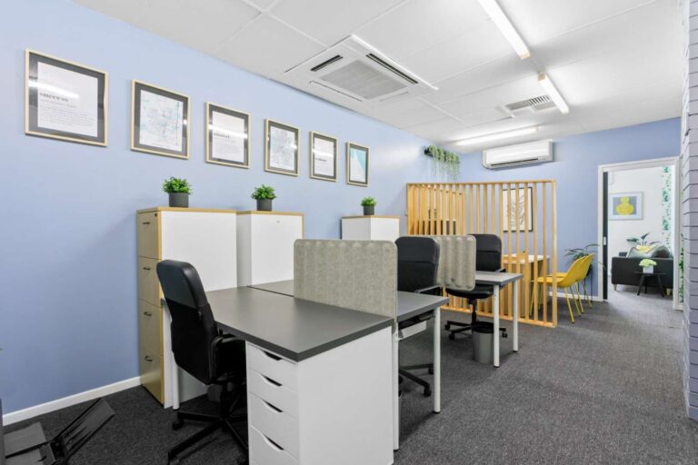 Modern office space with desks, black and grey chairs, light blue walls, framed pictures, plants on cupboards, and a partition separating workstations from a small meeting area.