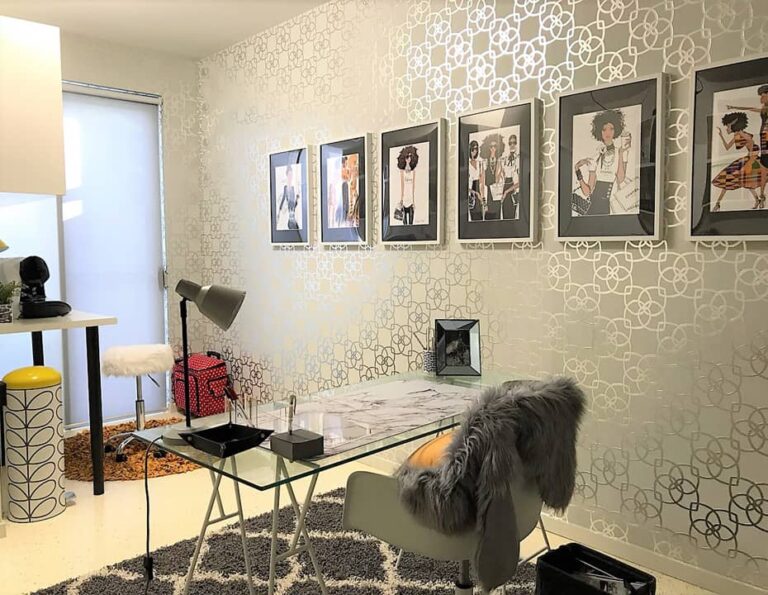 Modern home office with glass desk, white chair with fur throw, patterned rug, framed fashion photos on the wall, and a small bar area with two stools.
