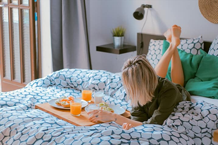 A person lies on a bed with geometric-patterned sheets, eating breakfast from a tray with juice, snacks, and salad. The room has a plant, a lamp, and cushions.