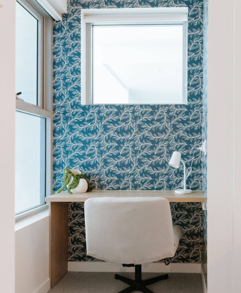 A small home office with a desk, white chair, potted plant, and lamp against blue leafy-patterned wallpaper. Two windows provide natural light.