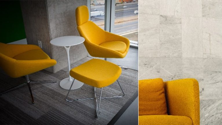 Yellow lounge chair with matching ottoman and a white round side table on a carpeted floor near a large window; partial view of another yellow chair against a grey tiled wall.
