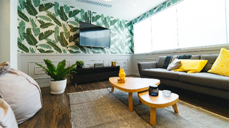 Modern living room with patterned leafy wallpaper, wall-mounted TV, grey sofa with yellow cushions, two wooden coffee tables, a plant, and large windows letting in natural light.