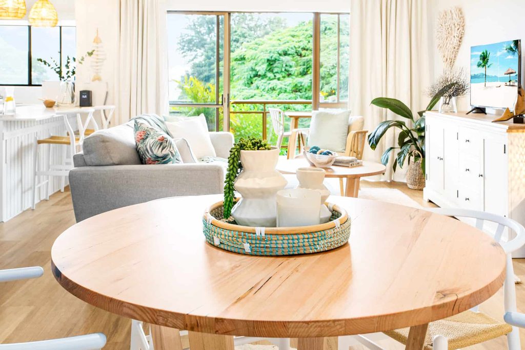 Bright living and dining area with wooden table, modern decor, sofas, large windows, and a view of greenery outside.