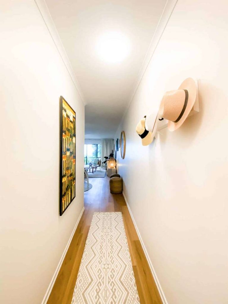 A narrow corridor with a patterned runner, hats hanging on the right wall, a colourful painting on the left, and a living area visible at the end.