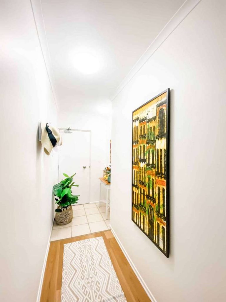 A narrow corridor with white walls, a patterned rug, a potted plant, hats on pegs, and a large painting of a building on the right wall.