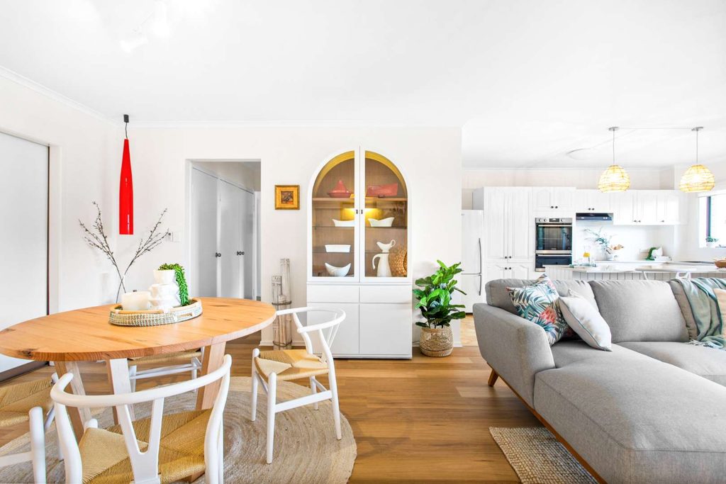 Bright, modern open-plan living and dining area with wooden floors, round table, white chairs, light grey sofa, built-in shelving, plants, and kitchen in the background.
