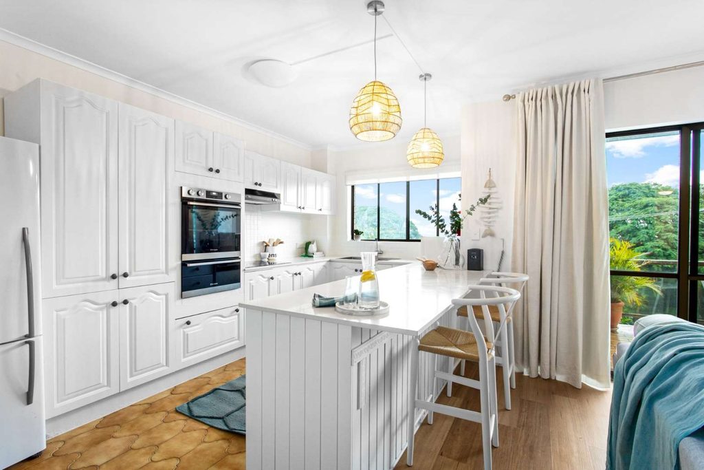 Bright modern kitchen with white cupboards, island with stools, pendant lights, large window with curtains, and natural light illuminating the space.