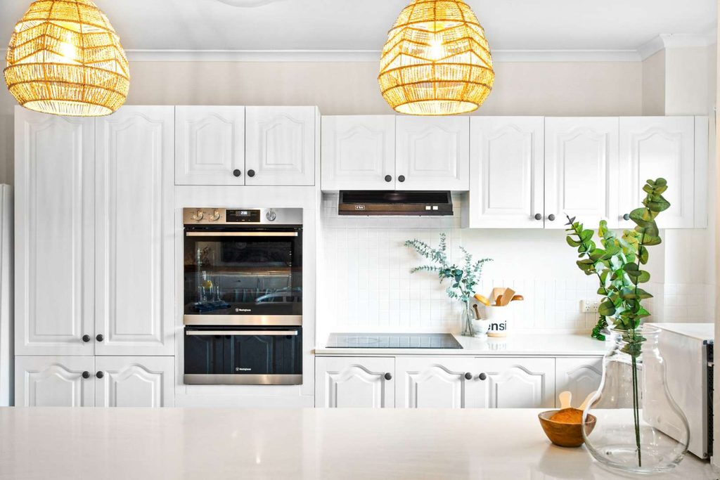 Modern white kitchen with built-in oven, hob, white cupboards, pendant lights, and a glass vase with green foliage on the worktop.