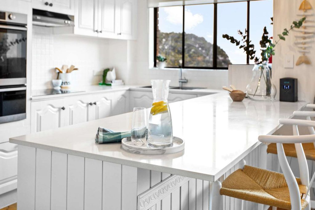 Bright white kitchen with a long island, two white chairs, a glass carafe with lemon water, and a window with a mountain view.