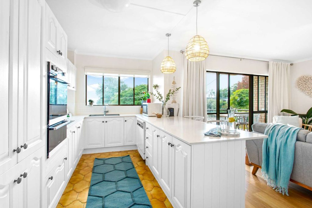 Bright kitchen with white cupboards, an island, pendant lights, blue hexagonal rug, large windows, and adjoining living area with a grey sofa and indoor plants.