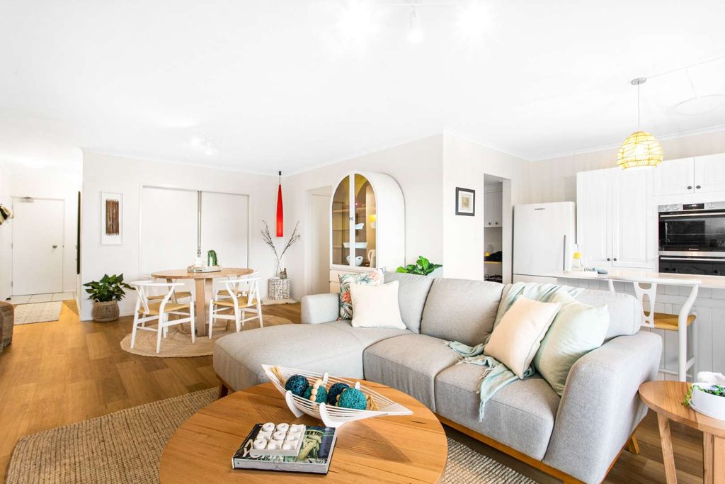 Bright, modern open-plan living room with a grey corner sofa, wooden coffee table, kitchen, and dining area, decorated in neutral tones with plants and coastal accents.