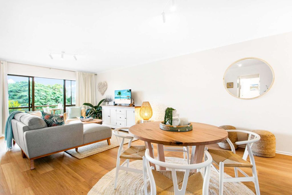 Bright living and dining area with wooden floors, round dining table, grey sofa, TV, indoor plants, and large window with greenery view.