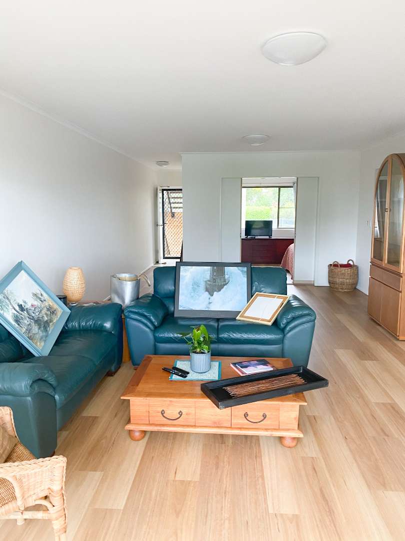 Living room with green leather sofas, wooden coffee table, framed art leaning on furniture, and a glass cabinet. Light wood floor and minimal decor. Bedroom visible in the background.