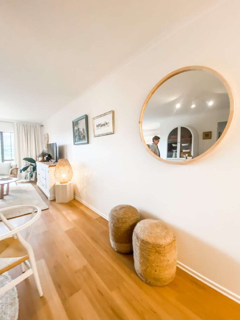 Bright living room with wooden floor, two woven stools, round wall mirror reflecting a person, art on the wall, TV stand, and lamp.