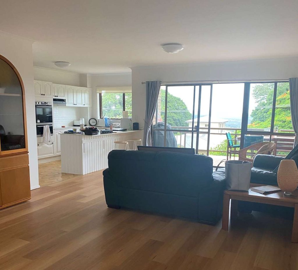 Open-plan living area with wooden flooring, black sofa, coffee table, and kitchen; large windows reveal a patio and greenery outside.