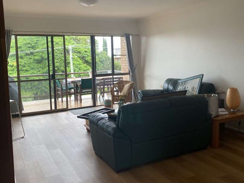 A living room with leather settees, a glass coffee table, wooden floors, and large windows leading to a balcony with outdoor seating and greenery outside.