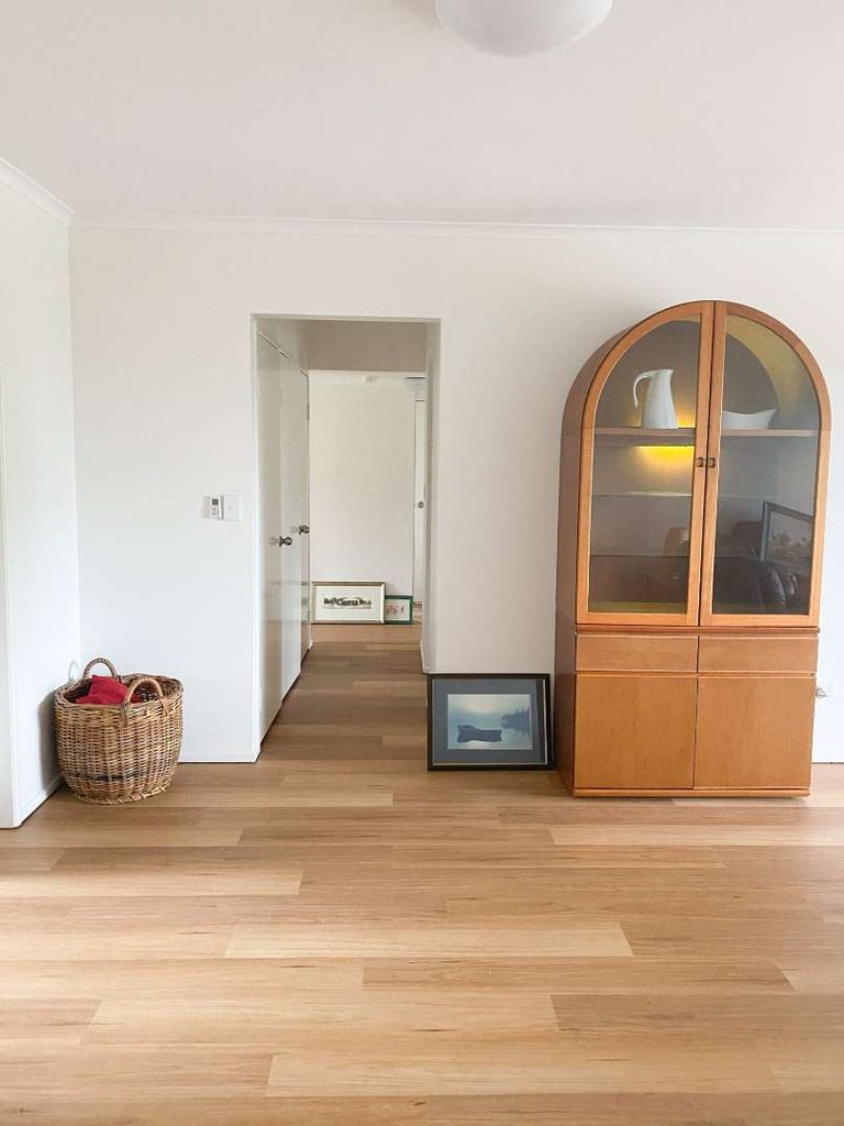 A minimalist room with light wood flooring, a brown cupboard with glass doors, a basket, and framed pictures leaning against a white wall near an open doorway.