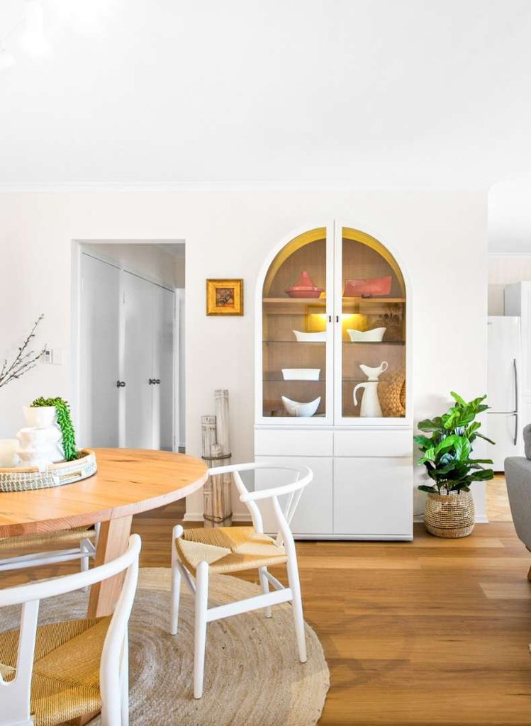 Bright dining area with a round wooden table, white chairs, a rug, and a white cupboard displaying dishes. A potted plant sits next to the cupboard on a wooden floor.