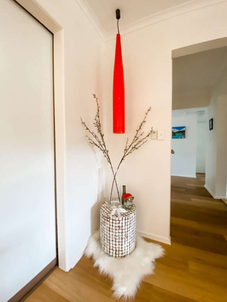 Tall red pendant light hangs above a decorative vase with branches and ornaments set on a white faux fur rug in a corner of a modern hallway with wooden flooring.