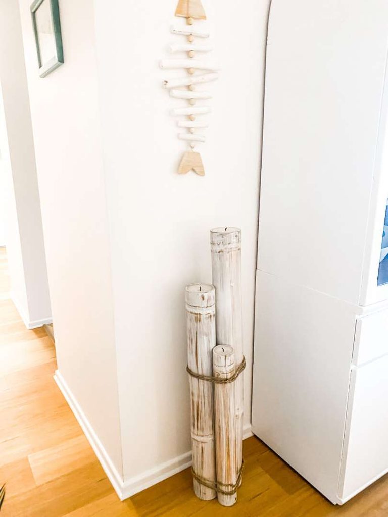 Three whitewashed bamboo poles are tied together and placed upright in a corner; above them hangs a minimalist wooden fish wall decoration on a white wall.