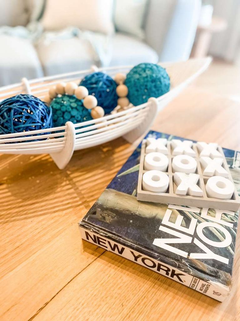 A white decorative bowl with blue spheres and beads sits on a wooden table next to a book titled “NEW YORK” with a noughts and crosses game on top.