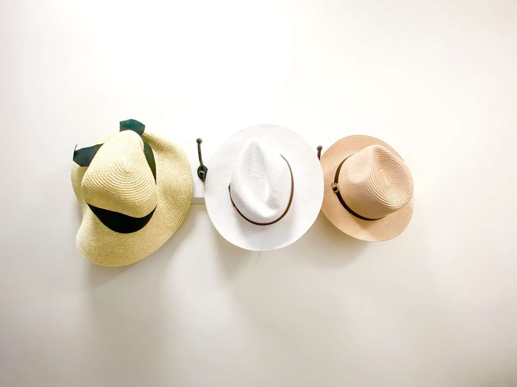Three hats—one light yellow, one white, and one tan—hang on wall hooks against a plain white background.