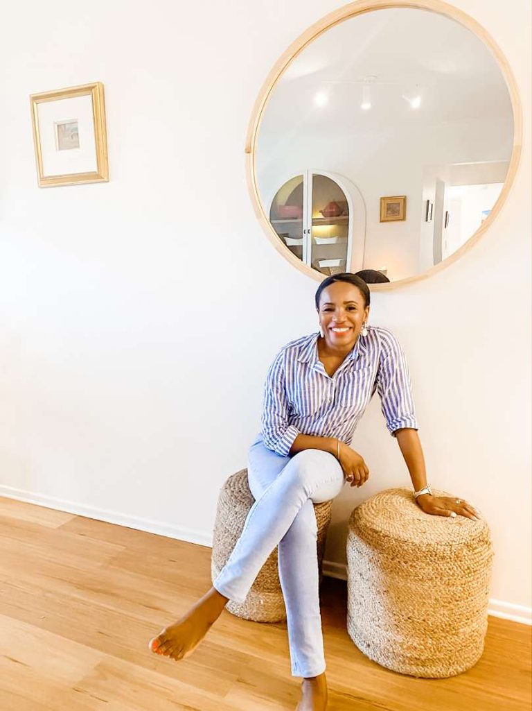 Woman sitting barefoot on a woven stool in a bright room with wooden floors, a round mirror, and framed art on the wall.