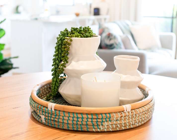A woven tray holds two white vases, one with a string of pearls plant, and a white candle on a wooden table in a bright, modern living space.