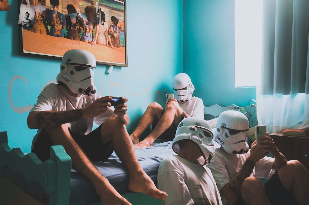 Four people wearing Stormtrooper helmets sit in a bedroom using their mobiles, with a beach-themed poster on the wall and sunlight coming through a window.