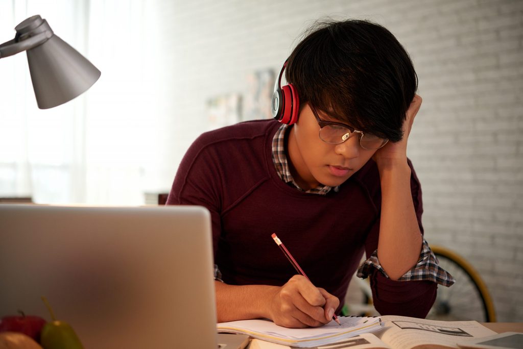 Young person wearing headphones and glasses writes in a notebook whilst looking at a laptop, seated at a desk with books and a desk lamp in a well-lit room.
