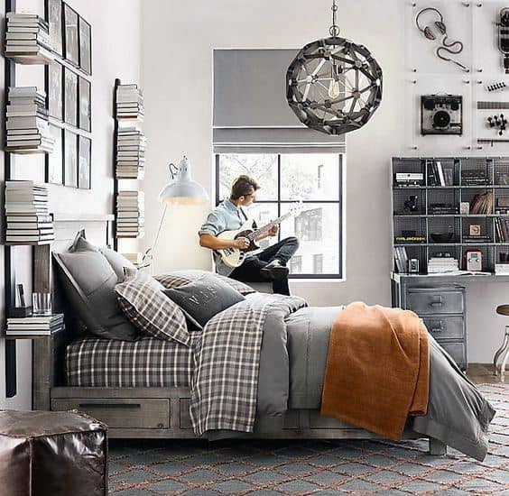 A neatly made bed in a modern bedroom with books and music décor; a person sits by the window playing an electric guitar.