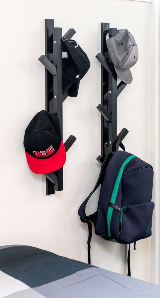 Two wall-mounted black racks holding three baseball caps and a blue rucksack, next to a bed with a grey and white blanket.